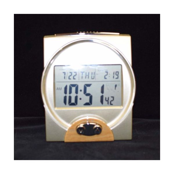 A square gold-colored clock with a large LCD display featuring current time, date, day, and alarm setting.